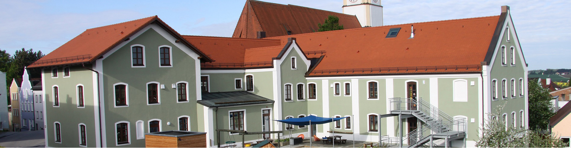 Das Foto zeigt den Kindergarten Haus Sonnenschein von der Georg-Brenninger-Straße aus. Man sieht das erweiterte Gebäude, das einen mittelgrünen Anstrich mit weißen Lisenen hat. Außerdem sieht man einen Teil des Außenspielbereichs.